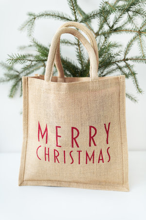 This is a photo of a burlap tote bag with Merry Christmas written in red on it, sitting in front of a small Christmas treeの写真素材