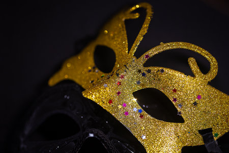 A close-up photo of two carnival masks, one gold and one black, on a black background. The gold mask is decorated with colorful rhinestonesの写真素材