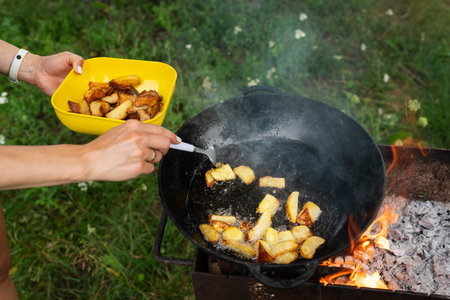 A man cooks potatoes in a wok over an open fire. Place the finished potatoes into a yellow bowl. The background is a grassy field. Outdoor picnicの写真素材