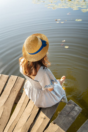 A young beautiful girl in a hat and a long dress sits with her back on the pier by the lake. Outdoor recreation conceptの写真素材