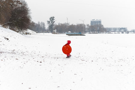 A small child in a bright red coat walks alone through a snowy landscape with a plastic sled, buildings visible in the distanceの写真素材