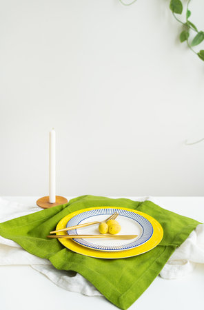 Minimalist table setting with green napkin, round plate with blue and yellow border, pair of fork and knife, white candle in background. Decor for the Easter tableの写真素材