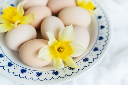 Fresh eggs and daffodils on a decorative plate, symbolizing spring or Easter. The plate is white with a blue pattern around the edge in the form of hearts and dotsの写真素材