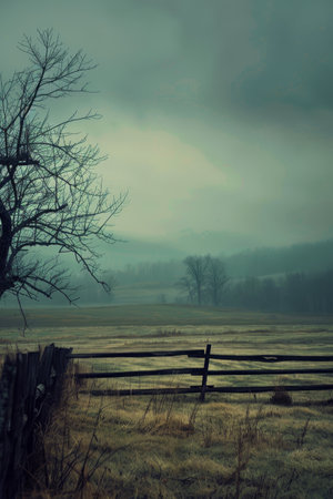 A moody rural landscape shrouded in fog, with a bare tree and a dilapidated fence, evoking the quiet of a late autumn day.の素材