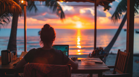 Woman working remotely on her laptop at a beach bar during a stunning sunsetの素材