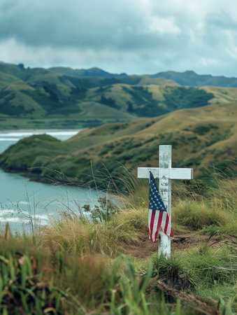 A weathered wooden cross adorned with an American flag stands on a grassy hillside, overlooking a picturesque lake and rugged mountain landscape, creating a solemn and reflective memorial settingの素材