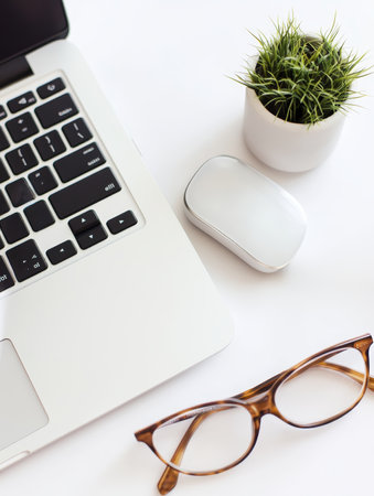 A clean, white desktop workspace featuring a laptop, wireless mouse, eyeglasses, and a small potted plant, embodying modern minimalist office design with copy space.の素材