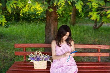 A young woman in a pink gingham dress sits on a red park bench, using her phone, with a basket of lupines beside her.の写真素材