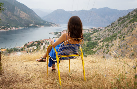 A woman in a blue dress relaxes on a yellow chair, enjoying a breathtaking view of a serene bay surrounded by mountains.の写真素材