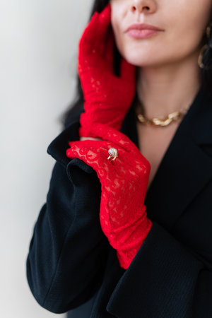 Elegant woman in red lace gloves and black blazer poses, touching face. Close-up highlights gold diamond ring over glove, gold necklace. Fashion, style, luxury accessories.の写真素材