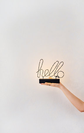 A woman's hand gently holds a decorative 'hello' sign with warm lights, creating a welcoming and...