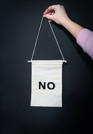 A hand holds a small beige banner with the word 'NO' boldly written in black, against a dark background.の写真素材