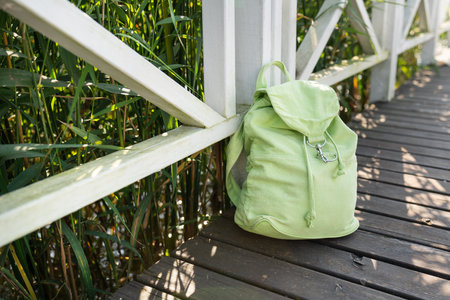 A light green canvas backpack rests against a white wooden fence beside a tranquil waterway.の写真素材
