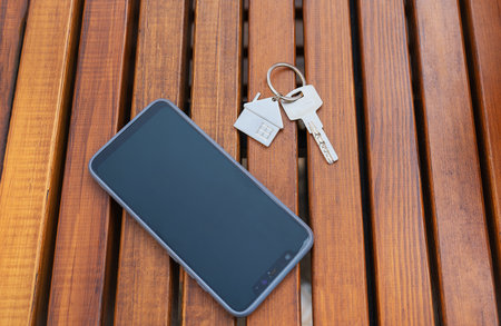 House keys with a key chain and a smartphone rest on a wooden surface, suggesting real estate or home ownership.の写真素材