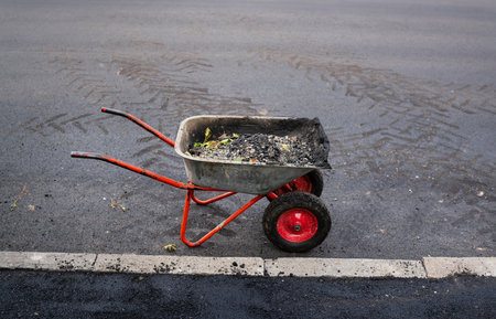 A wheelbarrow filled with asphalt and debris sits on freshly paved road next to a curb.の写真素材