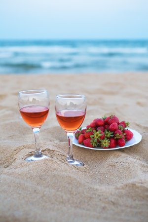 Two glasses of rose wine and a plate of fresh strawberries sit on a sandy beach, enjoying the ocean view.の写真素材