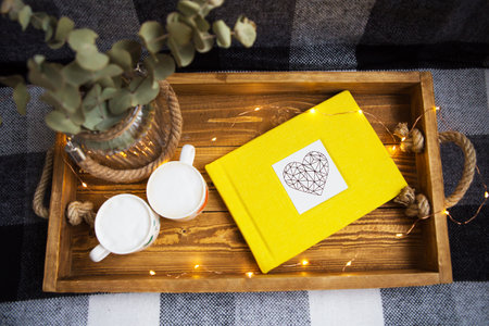 Cozy autumn scene with yellow book, mugs, and eucalyptus in a rustic wooden tray, decorated with...