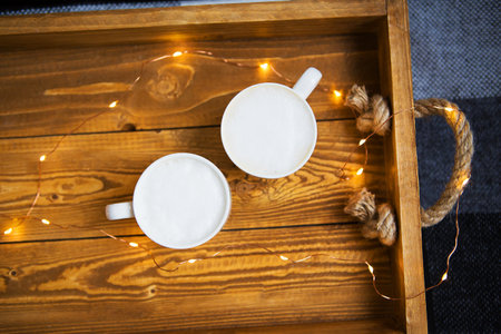 Two mugs of creamy latte art rest on a rustic wooden tray, adorned with warm fairy lights for a...