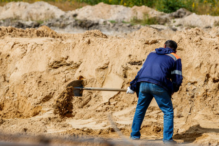 A man in a blue jacket and jeans diligently works with a shovel, digging sand at a construction site.の写真素材