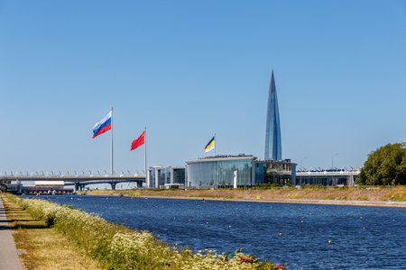 Lakhta Center and a rowing canal against a clear blue sky, flags, a bridge, and a shoreline are visible.の写真素材