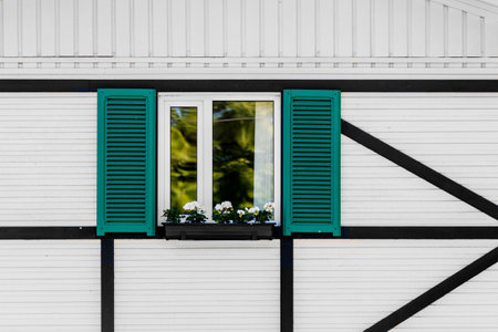A window with a white frame, green shutters, and a box with white flowers on the facade of a house with white siding and black beams.の写真素材