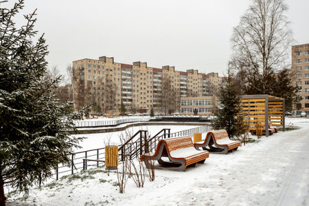 A snow-covered city park with benches and a fir tree against the backdrop of a typical Soviet residential building on a cloudy winter day.の写真素材