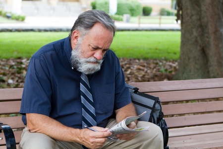 Unemployed middle aged man looks at advertisements for jobs in a newspaper while sitting on a park bench.の写真素材