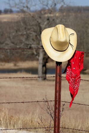 Yellow straw cowboy hat and red bandana hanging on the fence post of a rusty barbed wire fence with field and trees in the background.の写真素材