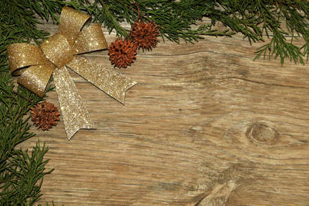 Green cedar branches, sycamore seed pods and a gold glitter bow frame a wood grain background with...