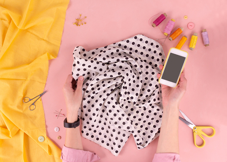 Women's hands hold the fabric in the workplace. The sewing process. Scissors, spools of thread, pins, phone. Fashionable concept. Flat layの写真素材