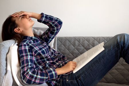 Distance learning online education and work. Tired girl sitting on the sofa, reading a book.の写真素材