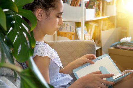 Beautiful teen girl reading a book sitting on the couch at home. Study, distance education,...