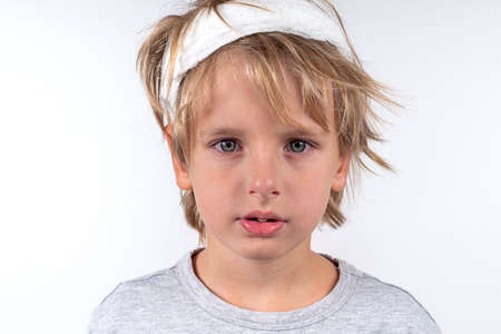 Portrait young caucasian cute boy blond hair with trauma injury and bandage head. Isolated on white background. Sick sad look boy. Red tired eyes from allergies.の写真素材