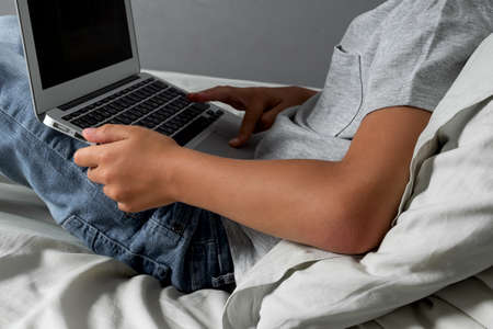 Little Caucasian blond boy with a head injury and bandage is sitting on the bed and using laptop. Recovery after incident. Remote communication, distance learning, technologies and gadgets in lifeの写真素材