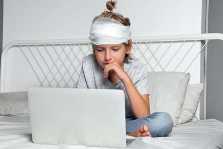 Little Caucasian blond boy with a head injury and bandage is sitting on the bed and using laptop. Recovery after incident. Remote communication, distance learning, technologies and gadgets in lifeの写真素材