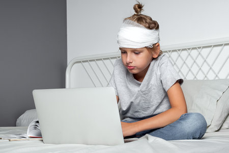 Little Caucasian blond boy with a head injury and bandage is sitting on the bed and using laptop. Recovery after incident. Remote communication, distance learning, technologies and gadgets in lifeの写真素材