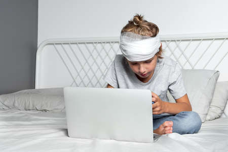 Little Caucasian blond boy with a head injury and bandage is sitting on the bed and using laptop. Recovery after incident. Remote communication, distance learning, technologies and gadgets in lifeの写真素材