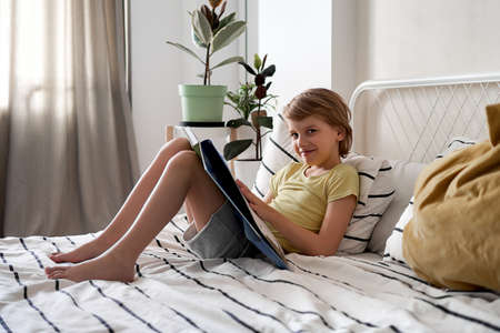A blond boy in a yellow T-shirt reads a book while lying on the bedの写真素材