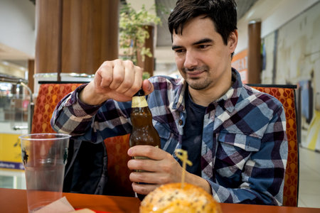 man eating fast food burger and drinking beer alone in the open area of a restaurant in a shopping...