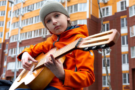 Portrait of caucasian blond boy in orange jacket playing acoustic guitar outdoors on autumn day