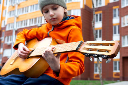 Portrait of caucasian blond boy in orange jacket playing acoustic guitar outdoors on autumn dayの写真素材