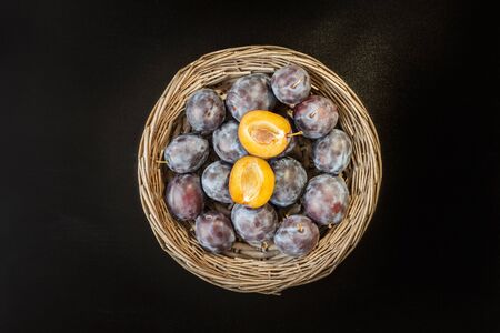 Fresh sweet plum in round rustic wicker basket on black chalkboard. Two half of plum yellow  core.の写真素材