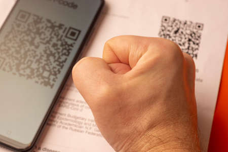 Fist of man next to smartphone with vaccination QR code and QR printed on paperのeditorial素材