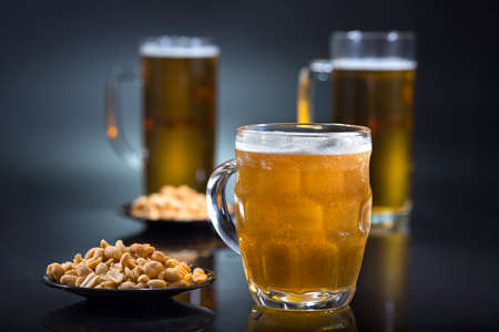 A glass of light beer and a plate of peanuts at the dark background.の写真素材