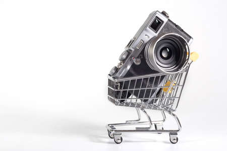 A camera in a small shopping cart at the white background. Selling photo technics, ideas of purchasesの写真素材