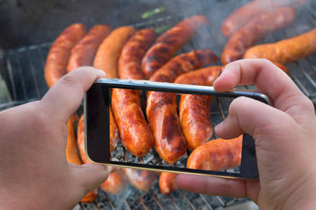 Food photography of baked sausages, meat. Photo of baked meat, shish kebabs with smartphone. Hands taking photo grilled fresh meat with smartphone.の写真素材