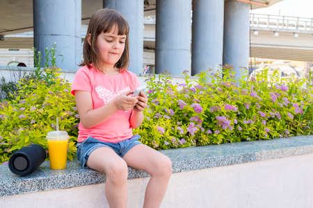 Cute preschool little girl, using smartphone, sitting on a parapet in city. A child holding smartphone playing a mobile game.の写真素材