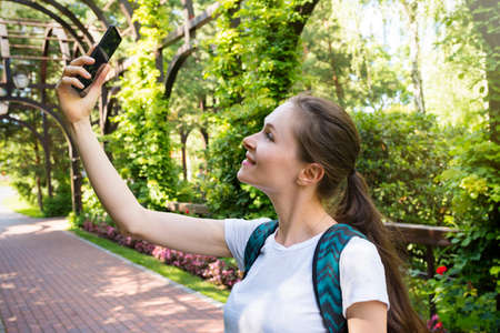 An outdoor portrait of a cheerful girl holding a smartphone in her hand, taking a selfie on the...