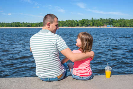 The father having embraced the daughter sit on the embankment against the background of the river in sunny summer day. Family day off.の写真素材