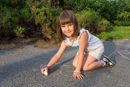 A little girl sits on the pavement and draws with crayons. Children's games in the open air.の写真素材